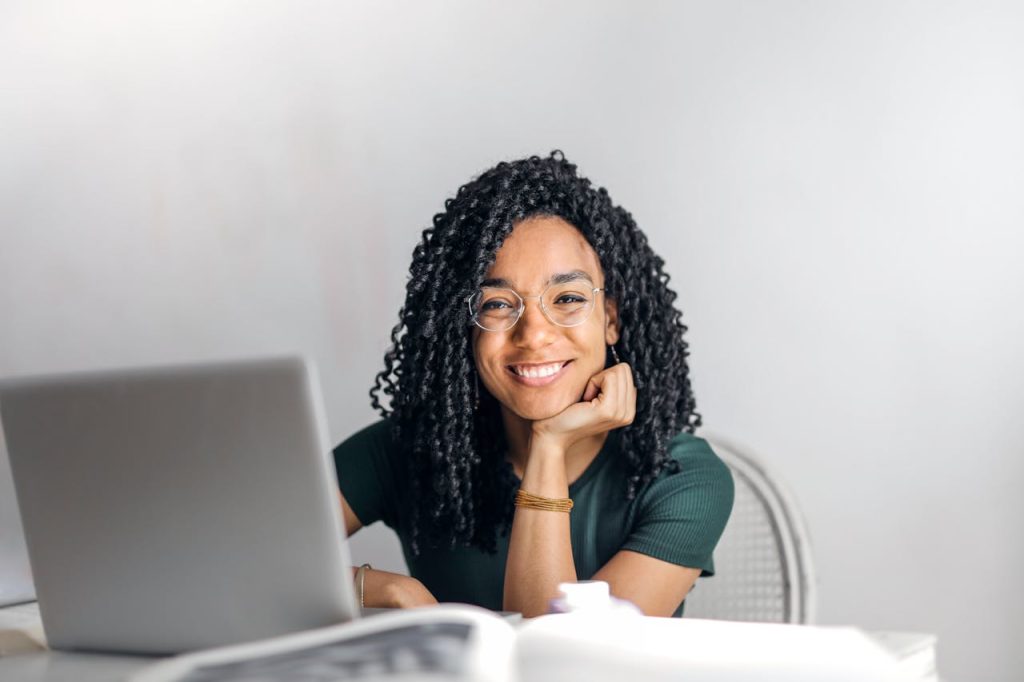 happy-ethnic-woman-sitting-at-table-with-laptop-3769021 Joyful businesswoman with curly hair smiling at camera while using laptop indoors.
