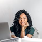 Joyful businesswoman with curly hair smiling at camera while using laptop indoors.